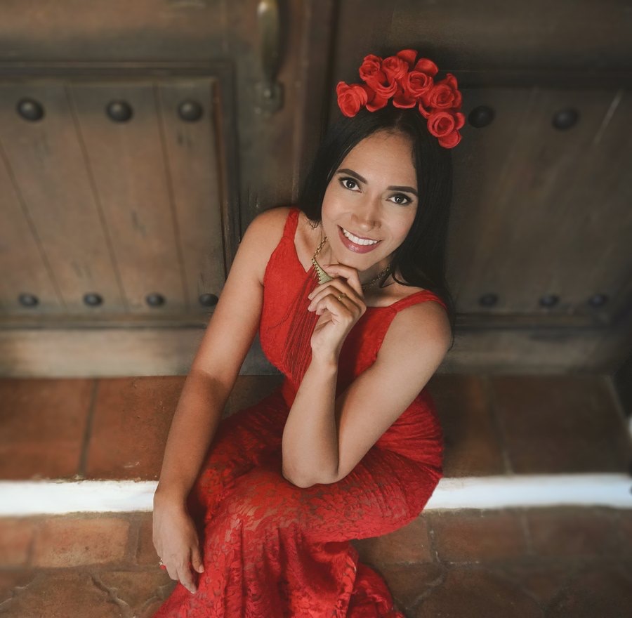 Rebeca Aldana con vestido rojo y corona de rosas frente a una puerta colonial